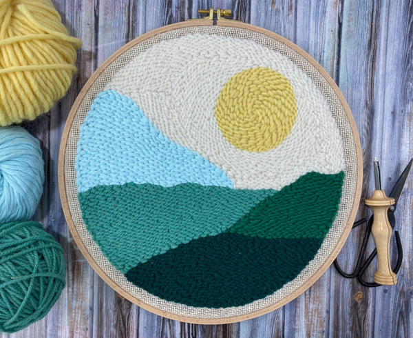 finished sun landscape punch needle wall hanging on a wooden table with a punch needle and yarn