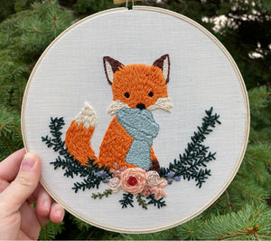Finished fox in a scarf punch needle design being held up in front of a fir tree