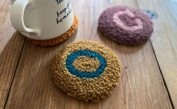 close up of "O" punch needle coaster, with two other coasters in the background.  One has a cup of coffee on it.