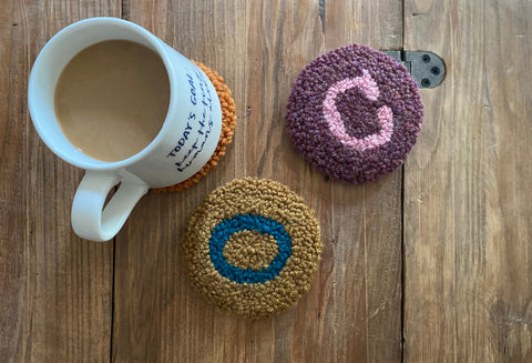 three punch needle coasters laying on a wooden table, one has a cup of coffee sitting on it