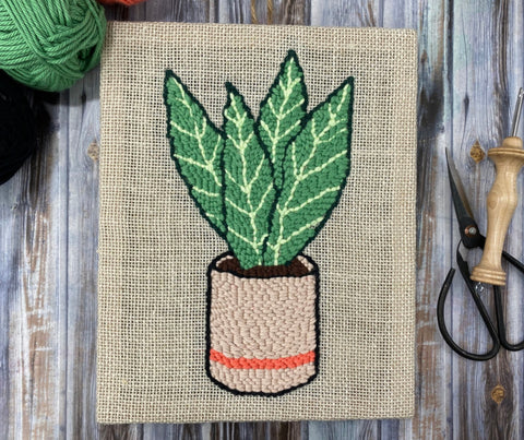 finished snake plant punch needle wall hanging on a wooden table with a punch needle and some yarn