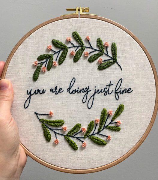 womans hand holding up "you are doing just fine" embroidered wall hanging